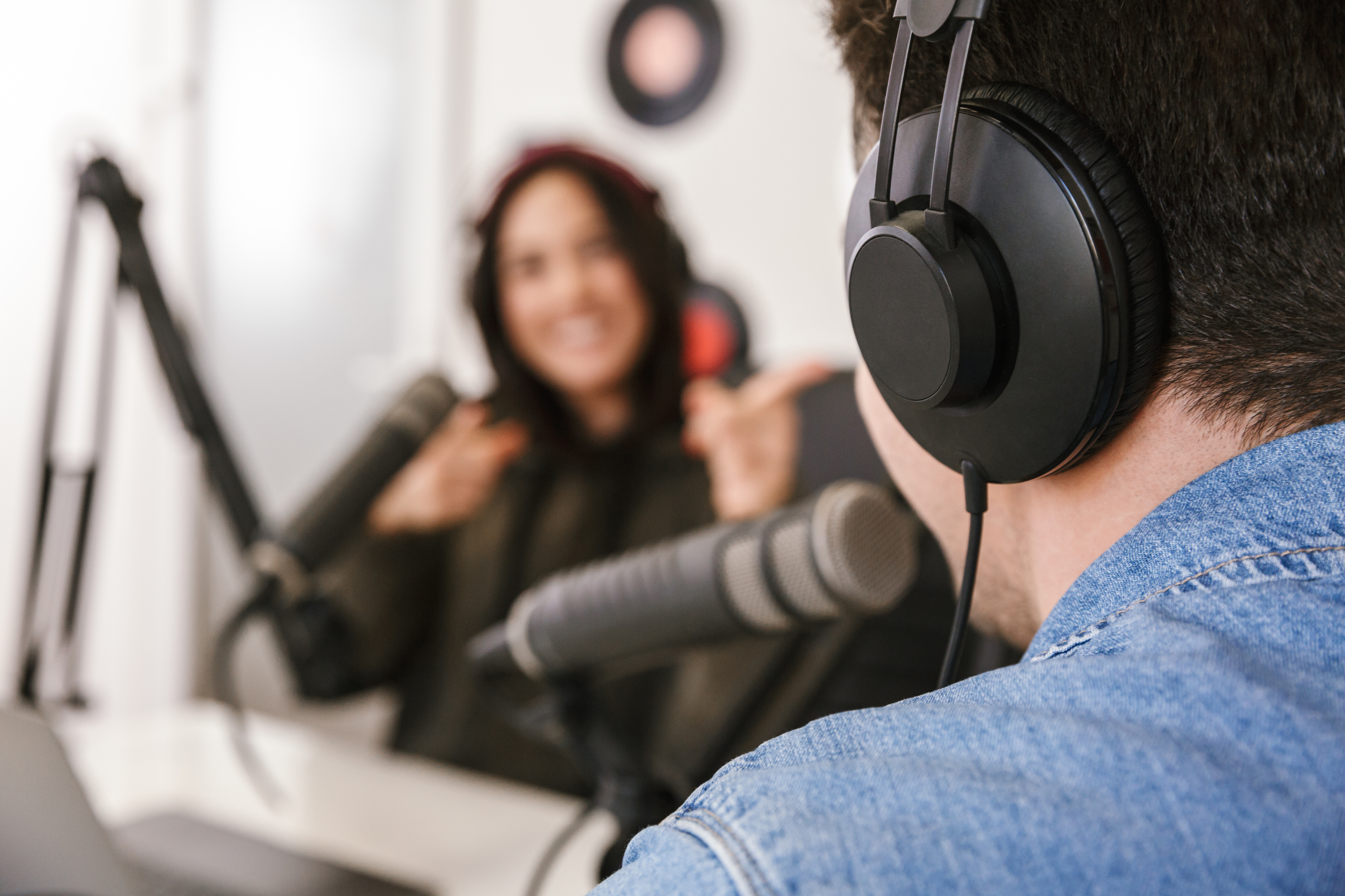 A man and a woman are recording a podcast. The man is seen from behind in the foreground. The woman is seen from the front and is laughing.