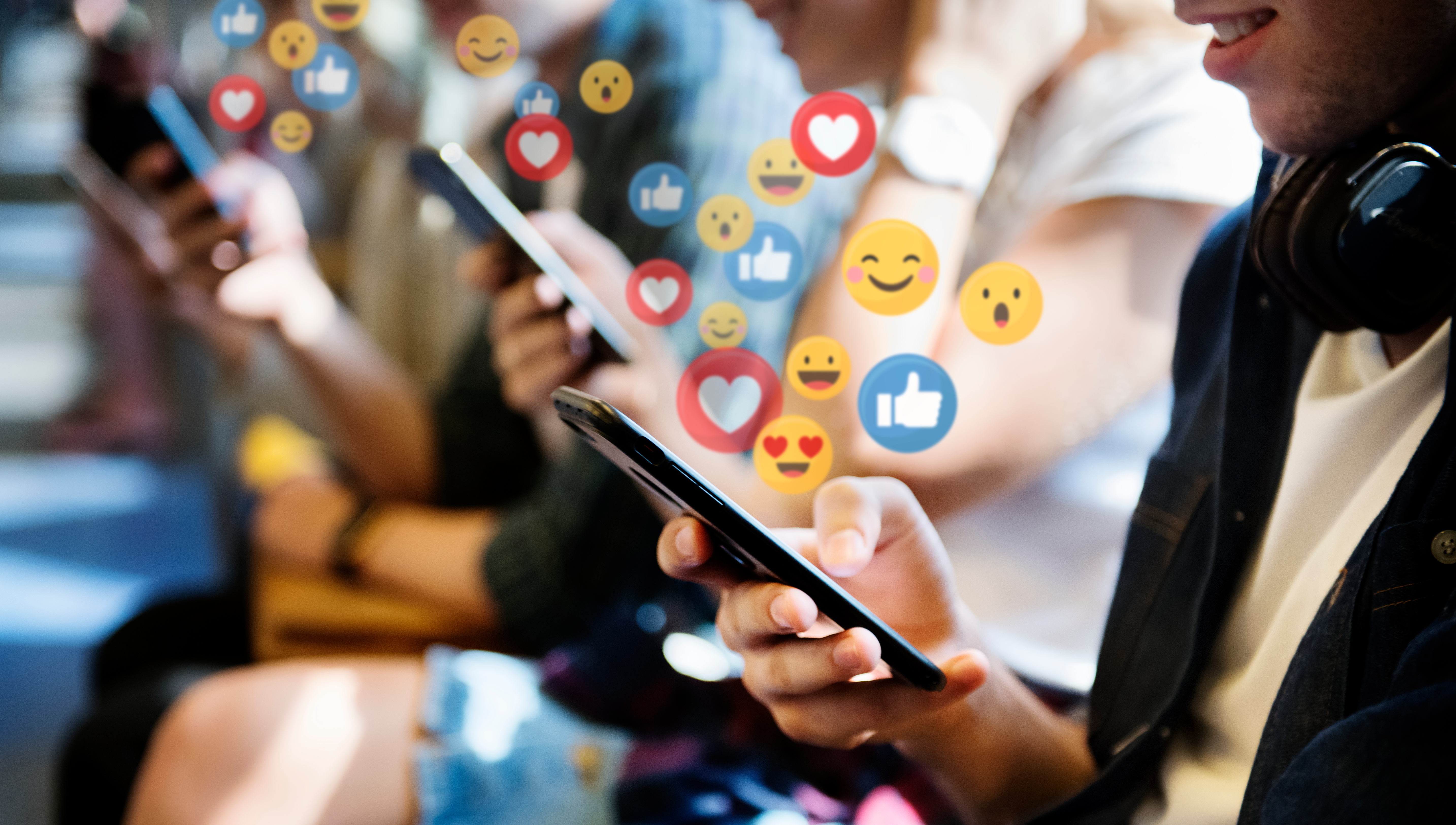 Several people sit next to each other in the subway and look at their cell phones. Social media icons such as emojis, thumbs up and hearts can be seen above the cell phones.