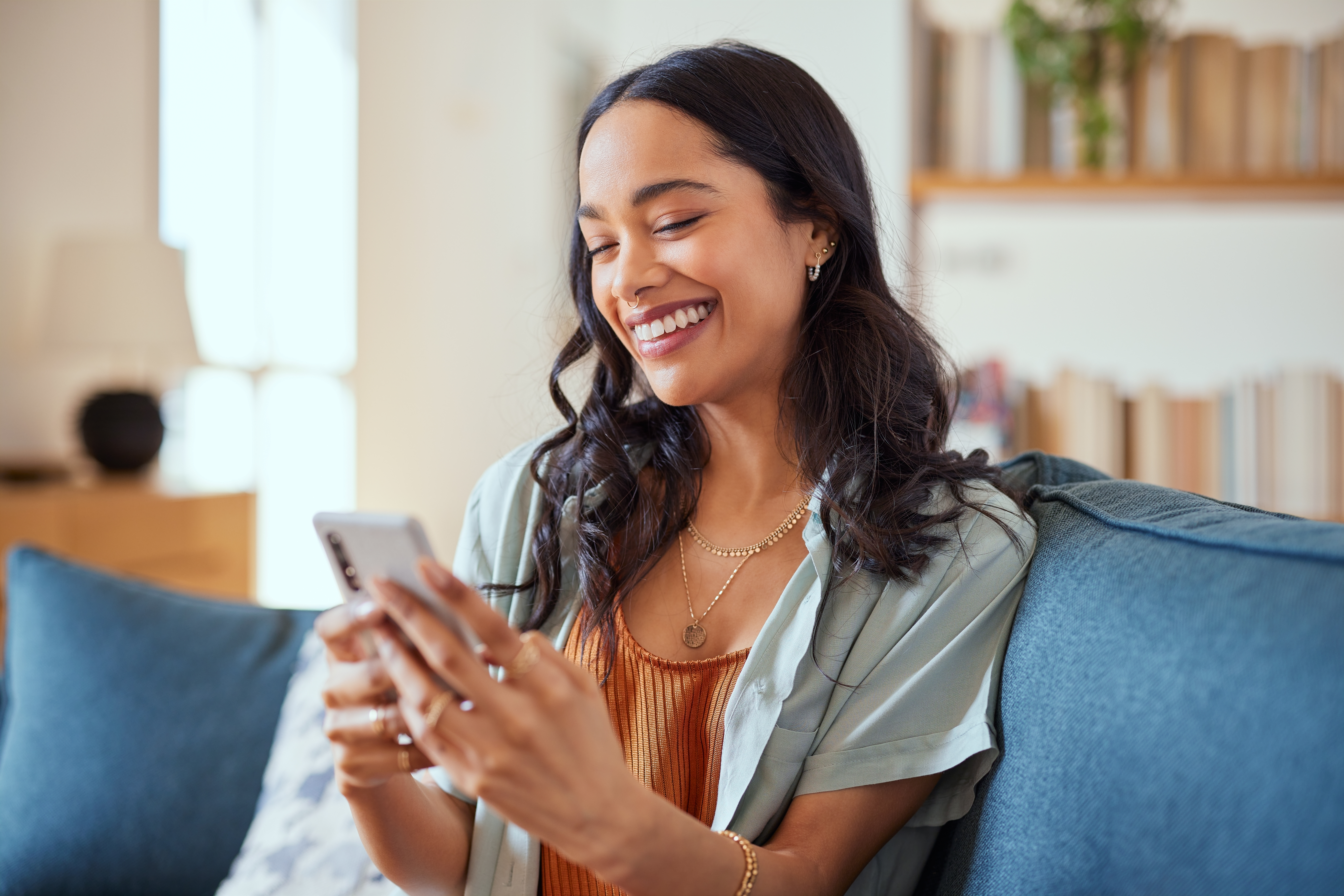 A woman sits on a couch and looks at her cell phone with a smile. Other furniture can be seen blurred in the background.