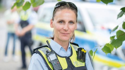 Portraitbild Polizistin Vanessa Göbel in Uniform mit blauer Schutzweste, Funkgerät, weißer Mütze unter dem Arm und Sonnenbrille auf dem Kopf vor einem verschwommenen Hintergrund; im Hintergrund ein Polizeifahrzeug und zwei Personen